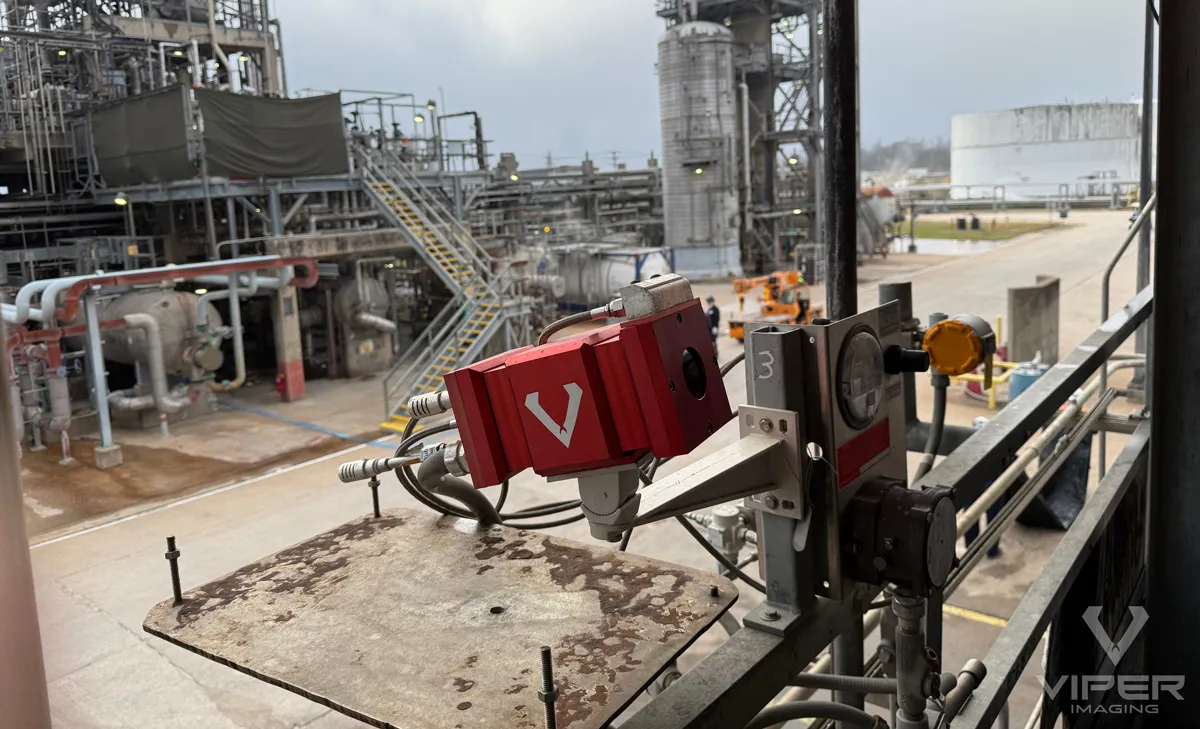 Red industrial camera mounted on metal bracket overlooking refinery piping, platforms, and storage tanks