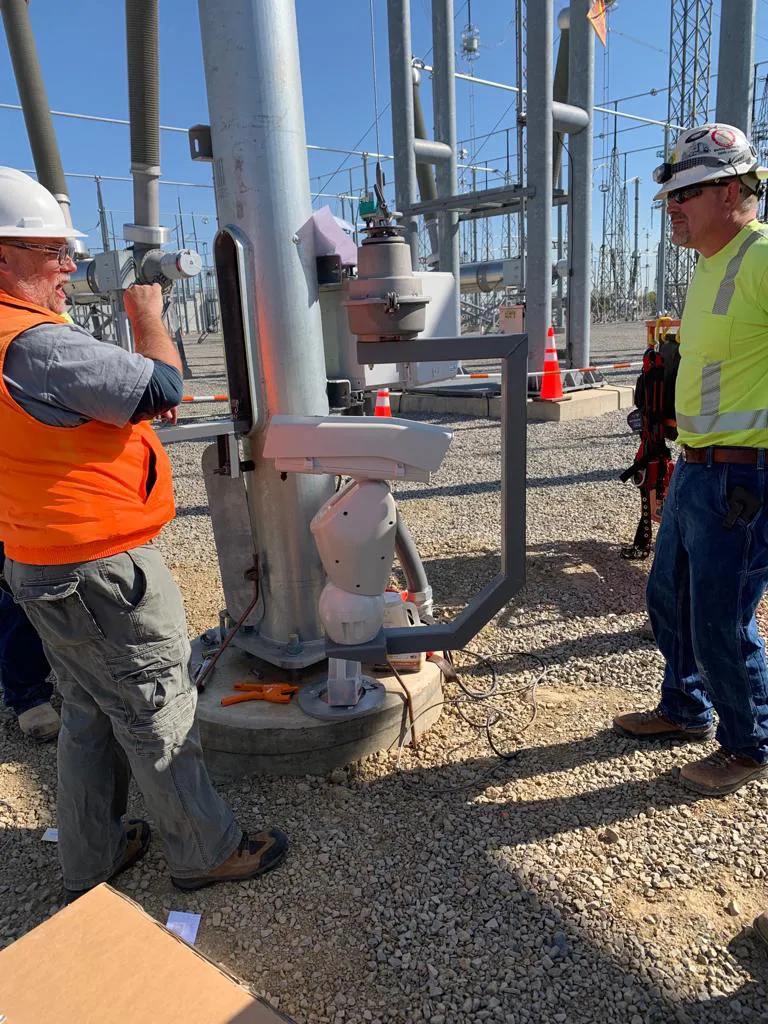 Two utility workers in hard hats and hi-vis gear inspecting electrical substation equipment at a steel pole